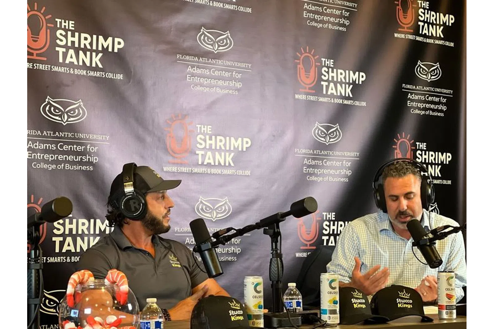 Two men wearing headphones speaking into microphones at The Shrimp Tank podcast backdrop with drinks and hats on the table
