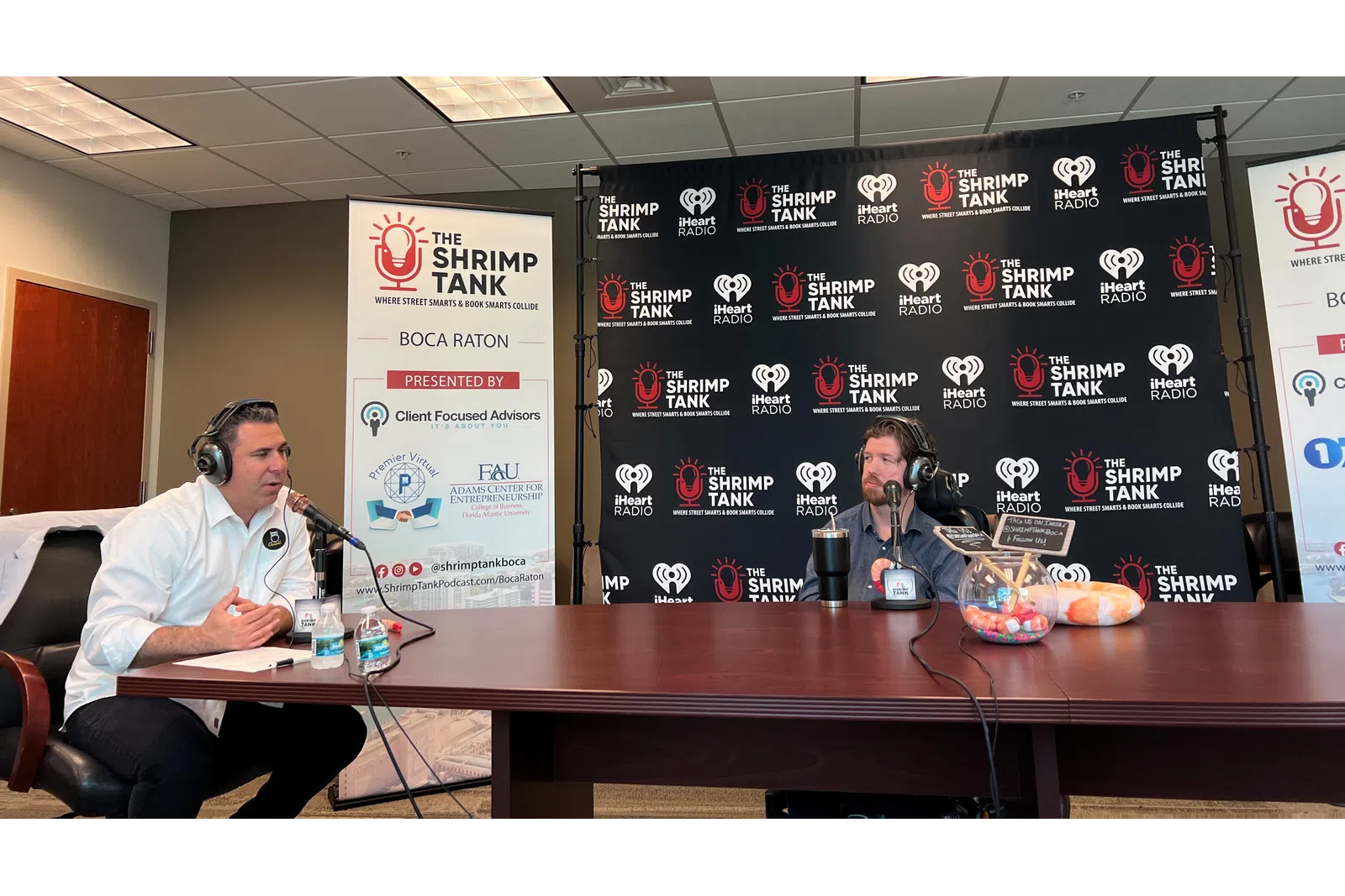 a couple of men sitting at a table with microphones and a sign