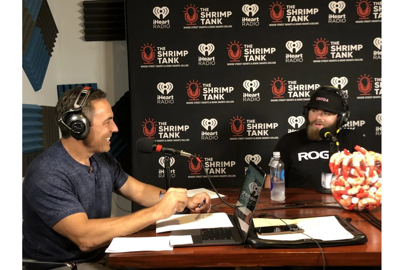 a couple of men sitting at a desk with microphones and a blackboard behind them