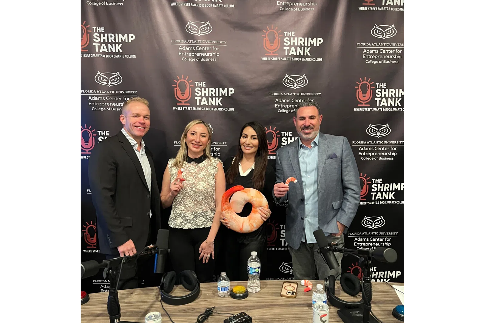 Four business professionals pose in front of The Shrimp Tank branded backdrop holding shrimp-themed items at a podcast setup.