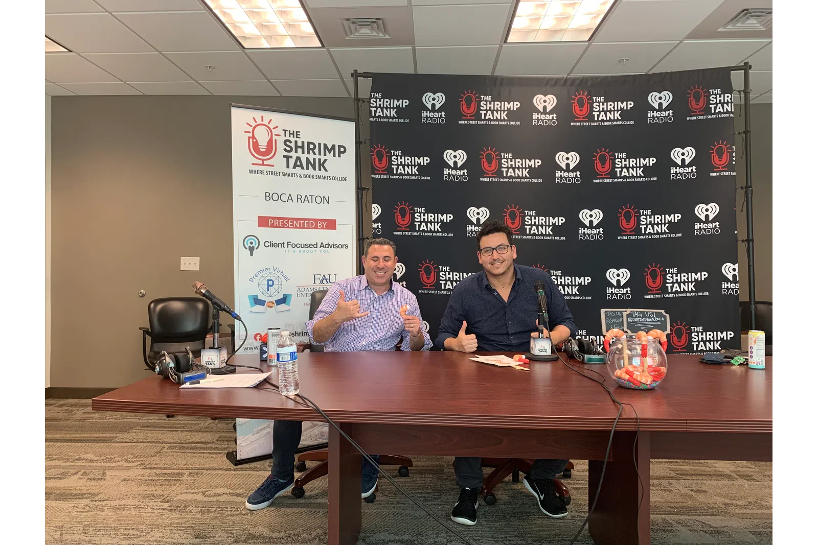 a couple of men sitting at a table with a sign behind them