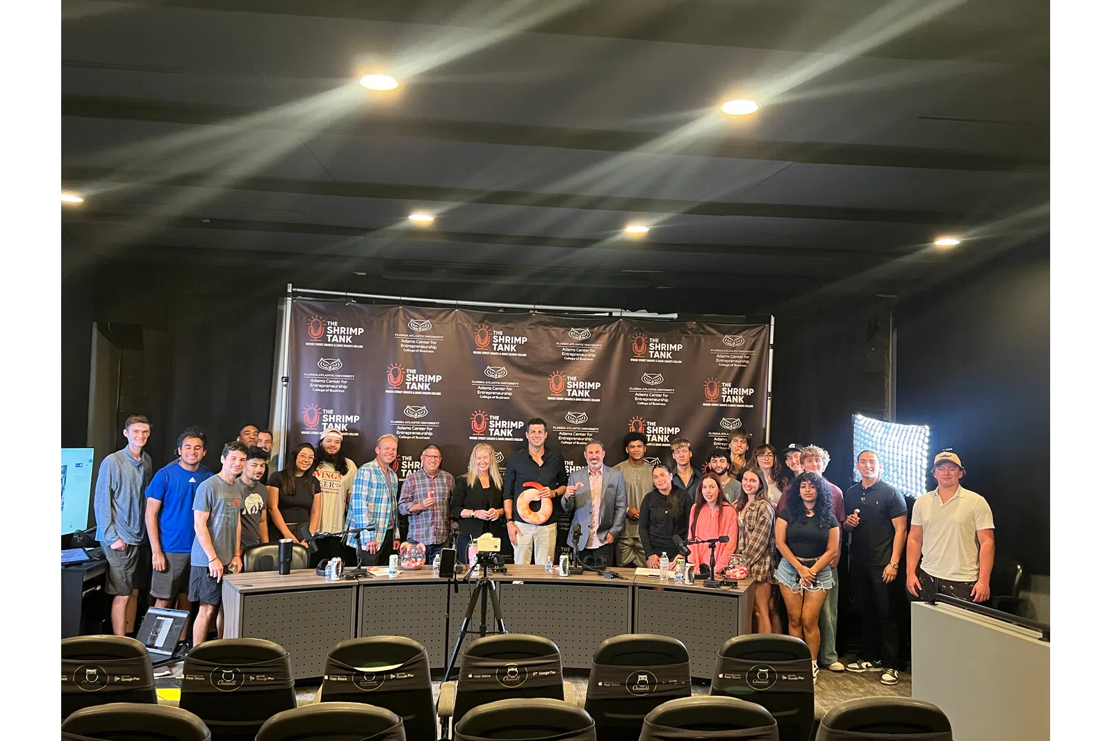 Group photo of diverse people standing behind a table with a Shrimp Tank backdrop and bright studio lights