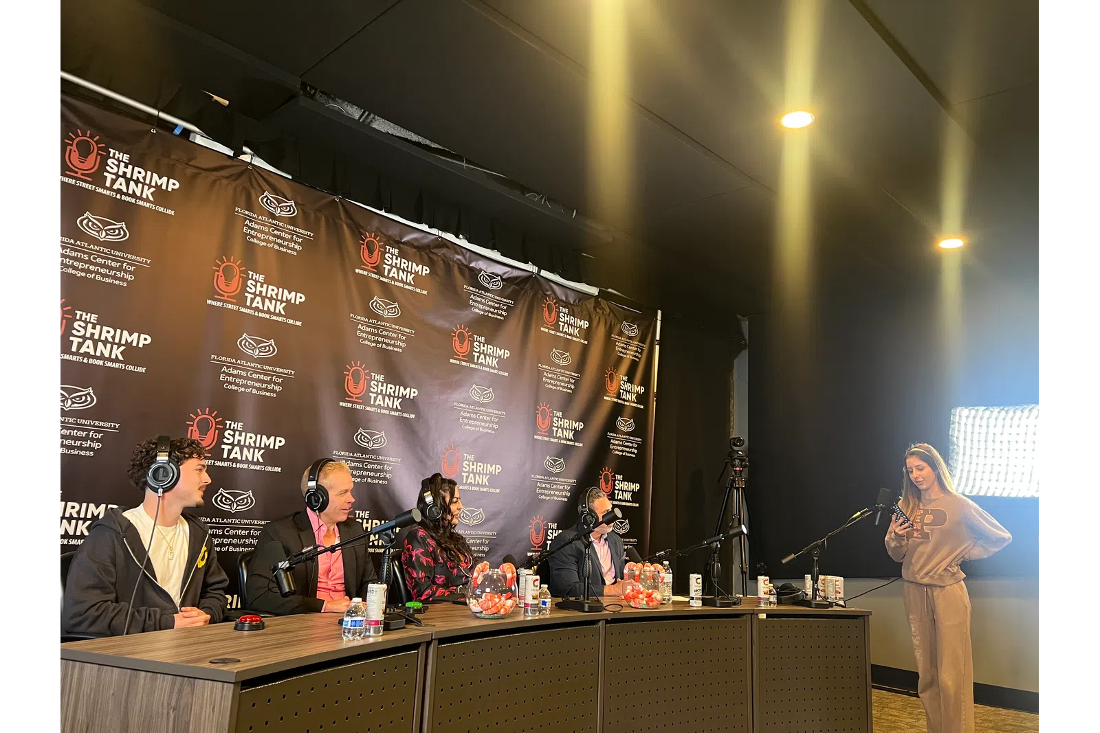 Four podcast hosts sit with microphones in front of a branded backdrop as a woman stands nearby with a smartphone and ring light.