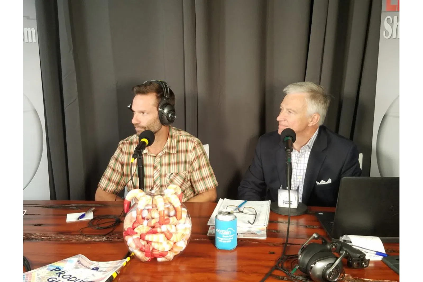a couple of men sitting at a table with microphones and food