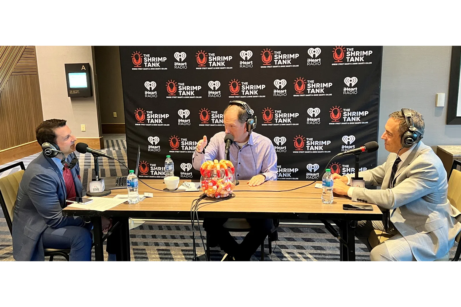 a group of men sitting at a table with microphones and a screen