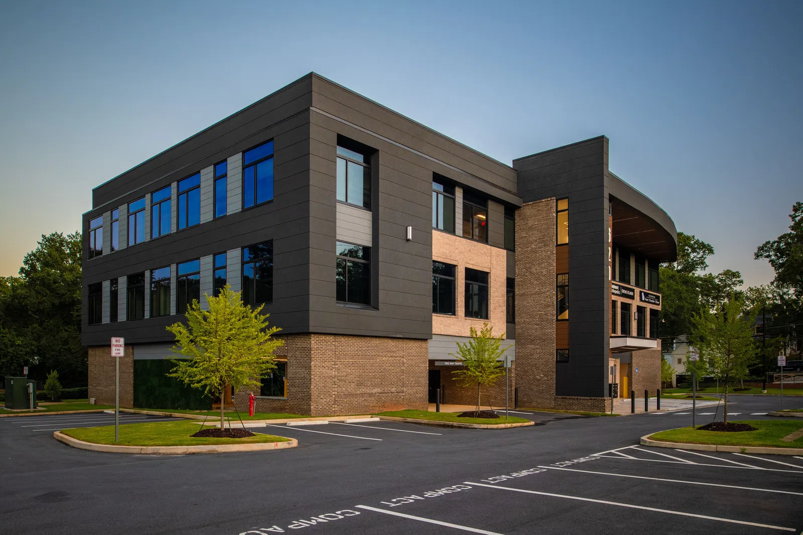 Modern two-story office building with black and brick exterior surrounded by trees and empty parking lot at dusk