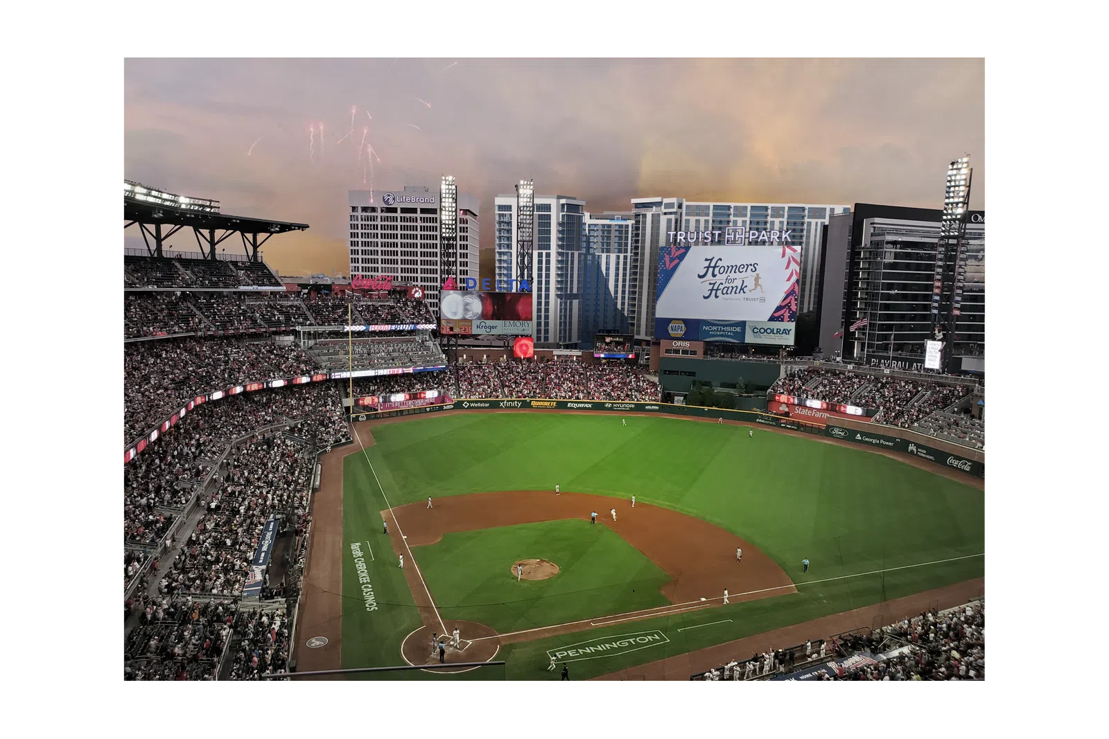 Twilight view of a packed baseball stadium with players on field and fireworks in sky.