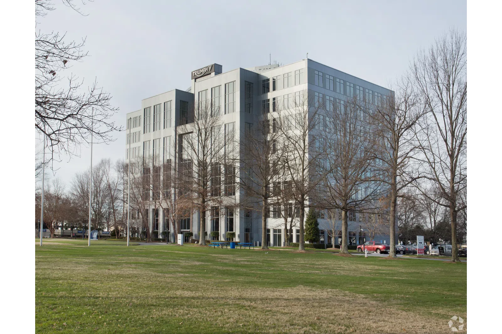Modern office building with glass windows behind leafless trees and a green lawn under cloudy sky