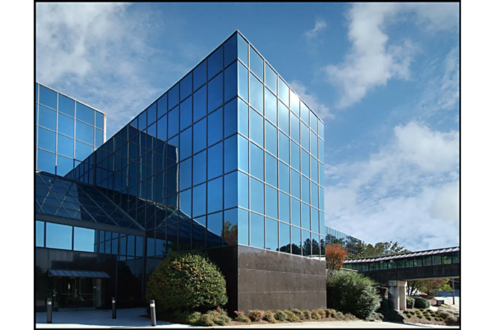 Modern glass office building reflecting blue sky with white clouds and surrounding greenery.