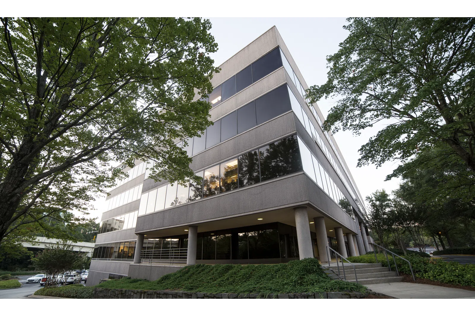 Modern office building surrounded by green trees with reflective windows and a sidewalk entrance.