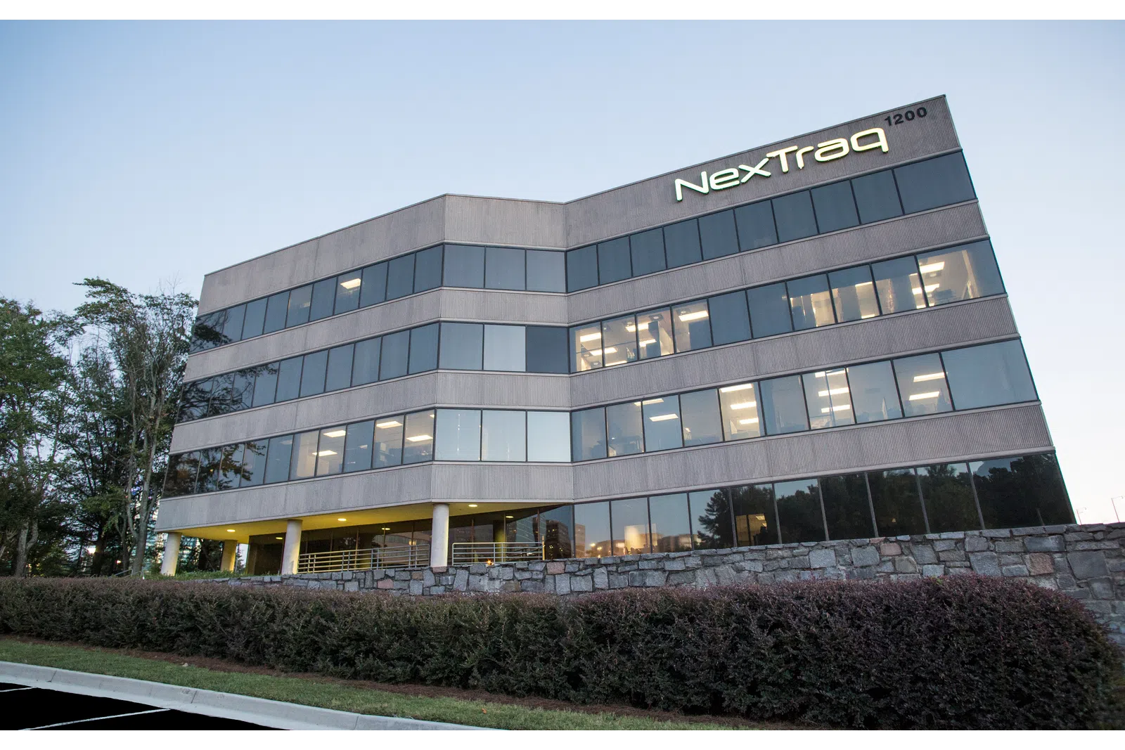 Modern office building with large windows and NextTraq logo at dusk surrounded by greenery and stone wall.