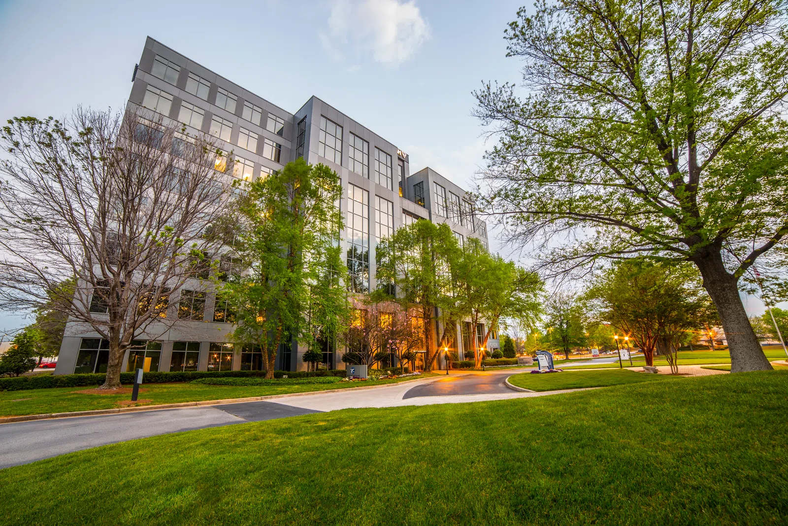 Modern office building with large glass windows surrounded by green trees and manicured lawn at dusk.
