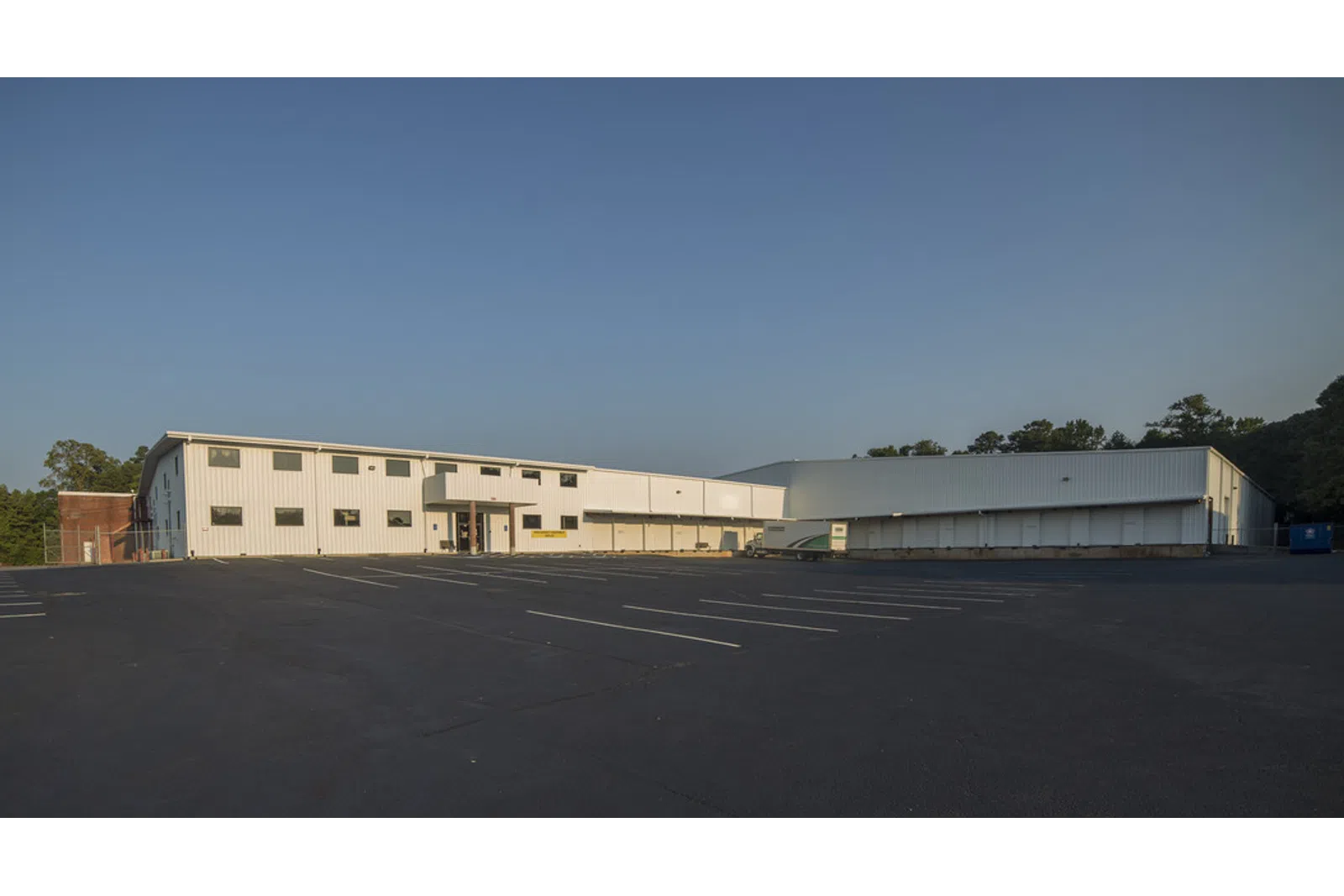 Large white industrial warehouse with empty parking lot under clear blue sky in daylight.
