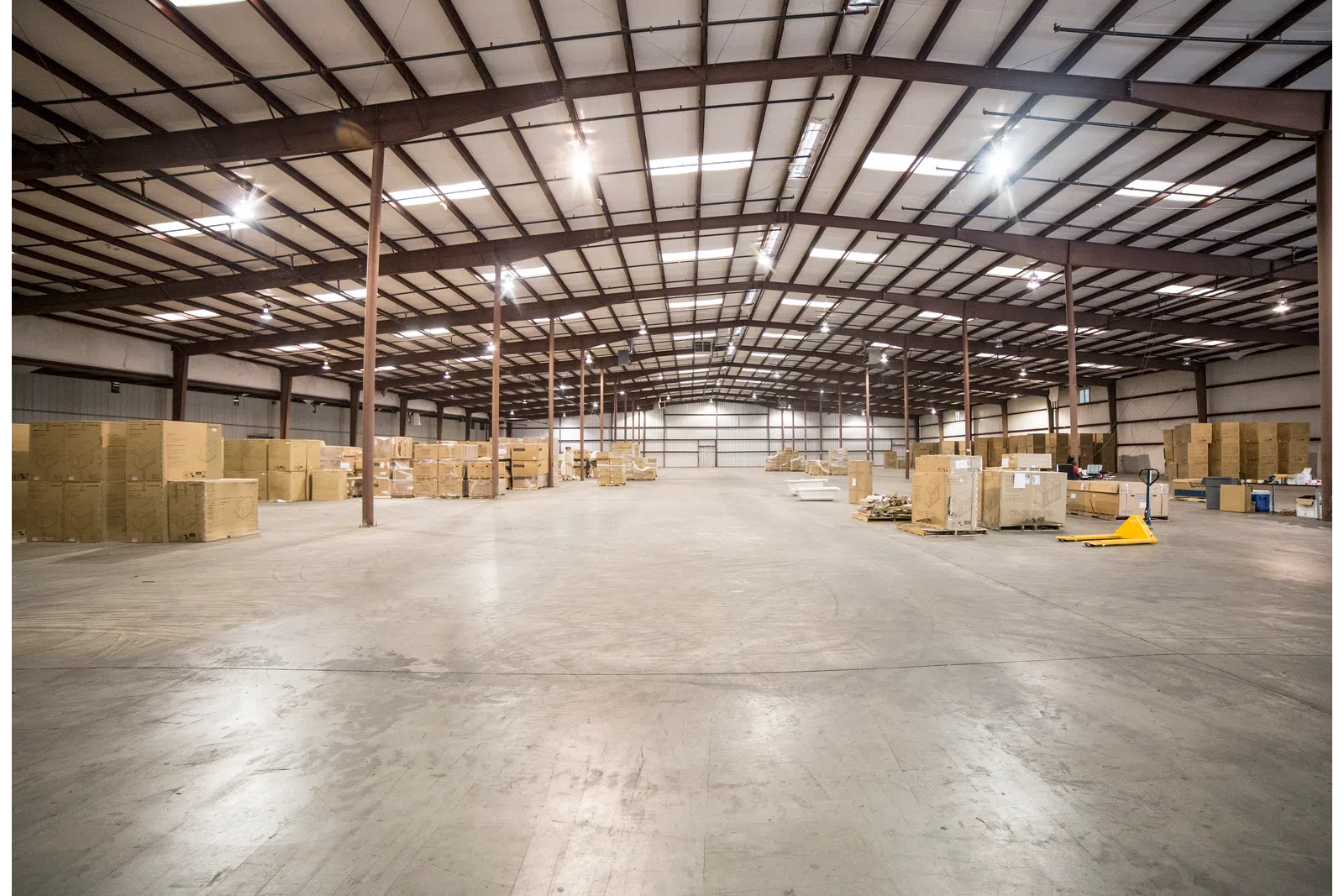 Spacious warehouse interior with concrete floor, metal beams, and stacks of cardboard boxes under bright lighting.