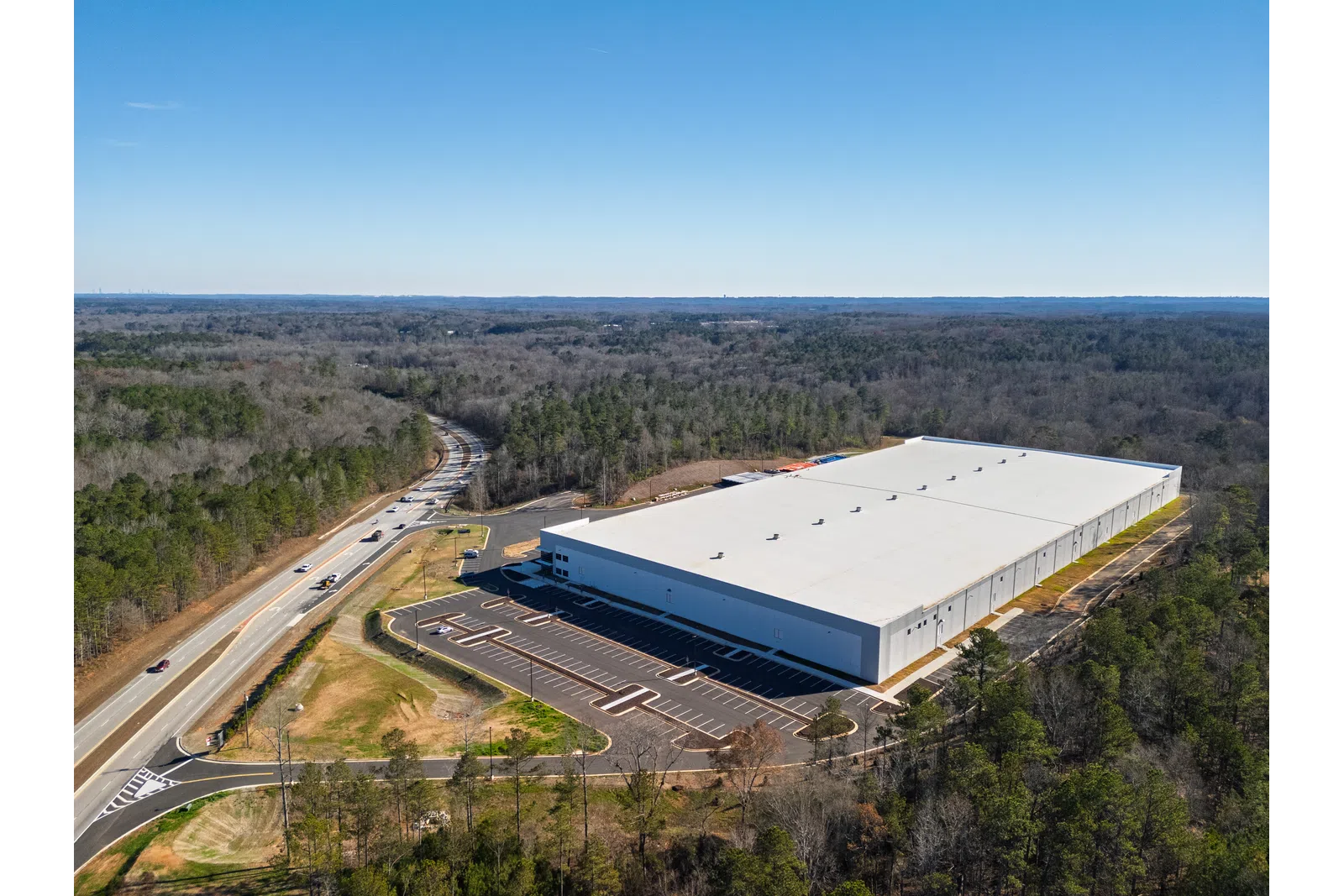 Aerial view of large white industrial warehouse surrounded by forest and adjacent to a road under clear sky