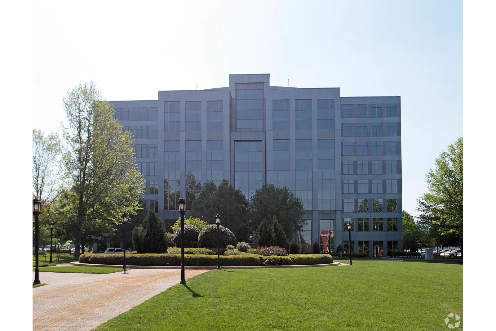 Modern glass office building with landscaped garden, trees, and lamp posts in the foreground on a sunny day