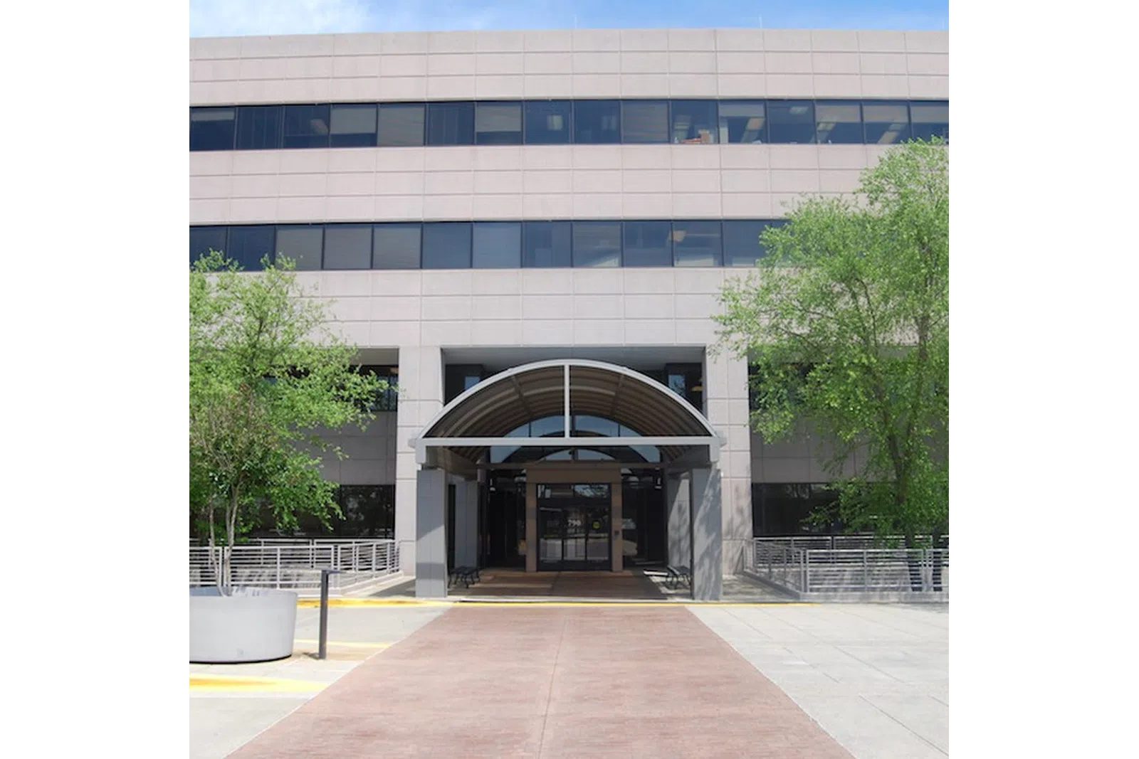 Modern office building entrance with arched canopy, surrounded by trees and concrete planters on a sunny day