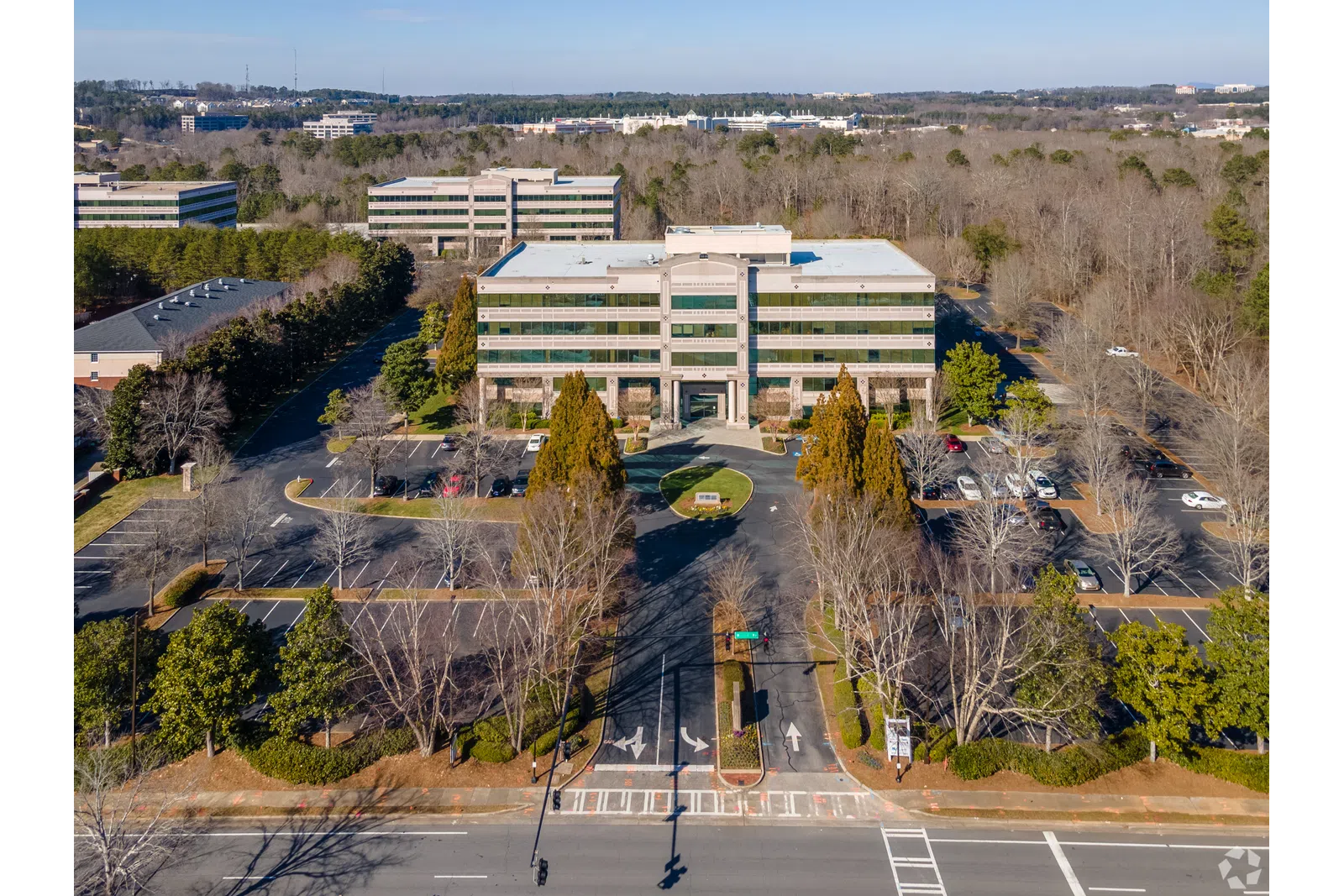 Aerial view of a modern office building surrounded by parking lots and trees in a suburban area under blue sky.