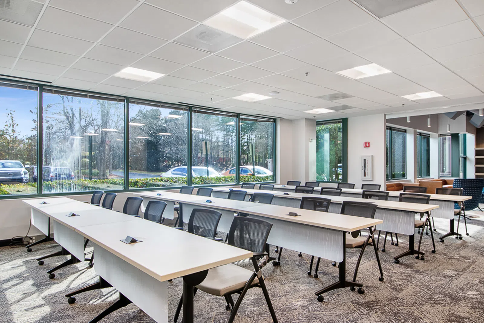 Bright modern conference room with large windows, rows of tables and black chairs, and carpeted floor.