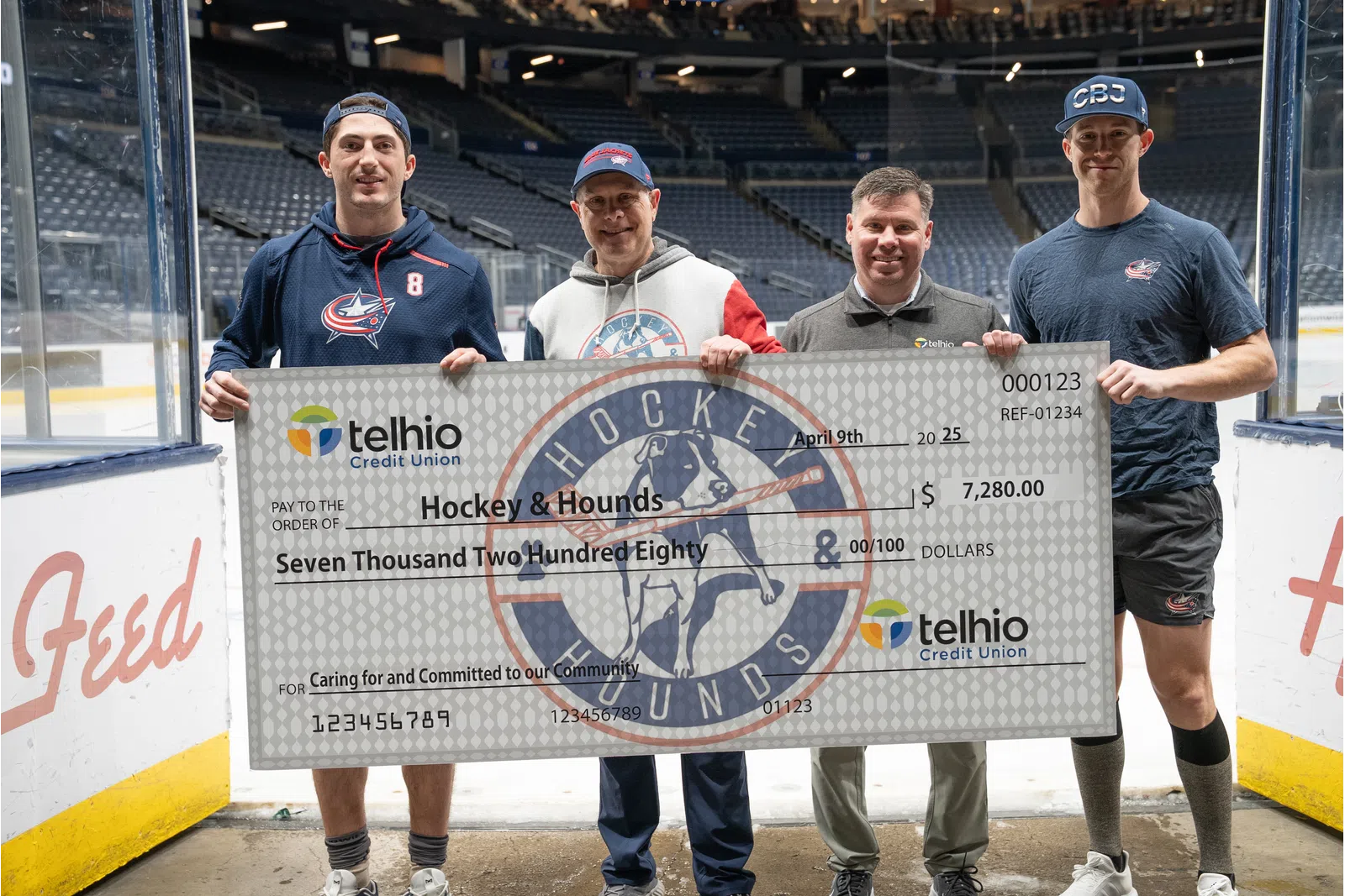 Four men on an ice hockey rink holding a large Telhio Credit Union check for $7,280 made out to Hockey & Hounds.