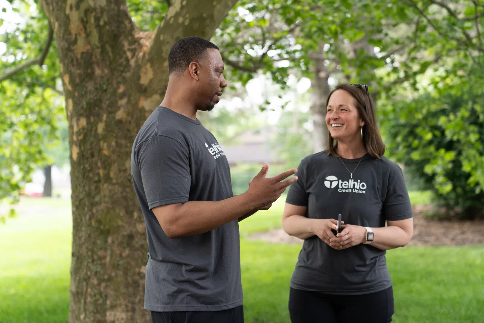 Two people wearing Telhio Credit Union shirts talking outdoors near a tree on a sunny day