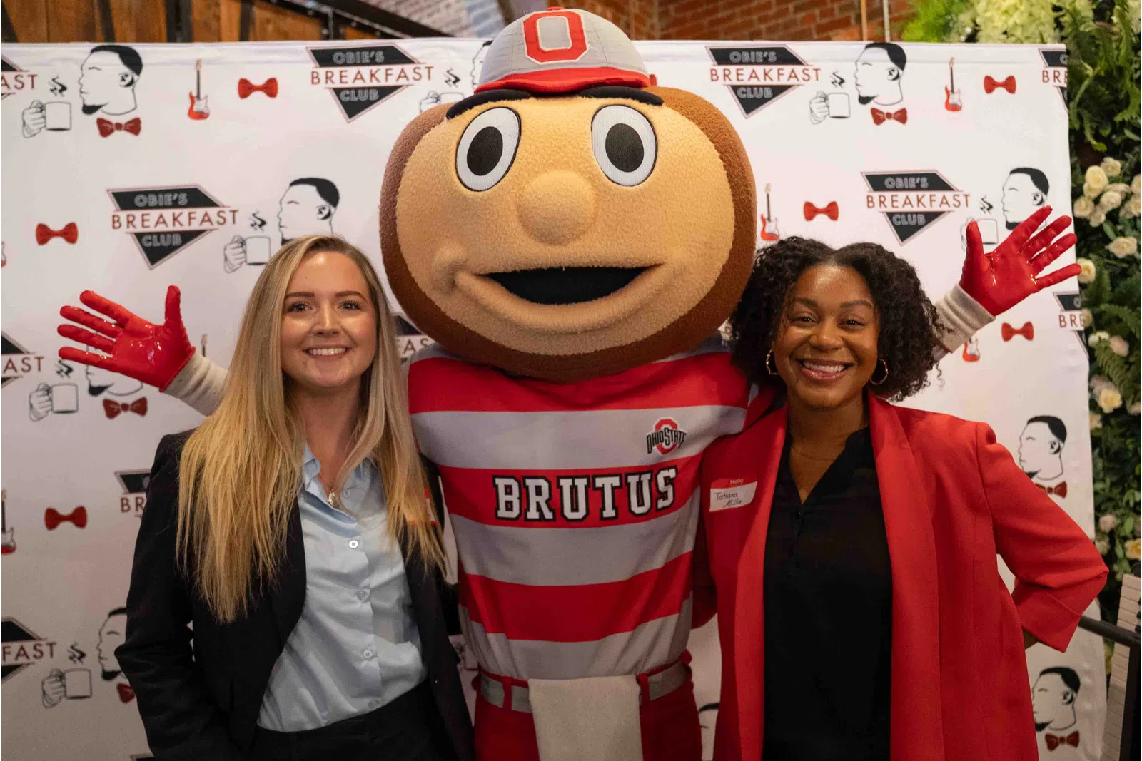 Two women smiling with Ohio State mascot Brutus Buckeye at Obie's Breakfast Club event backdrop.