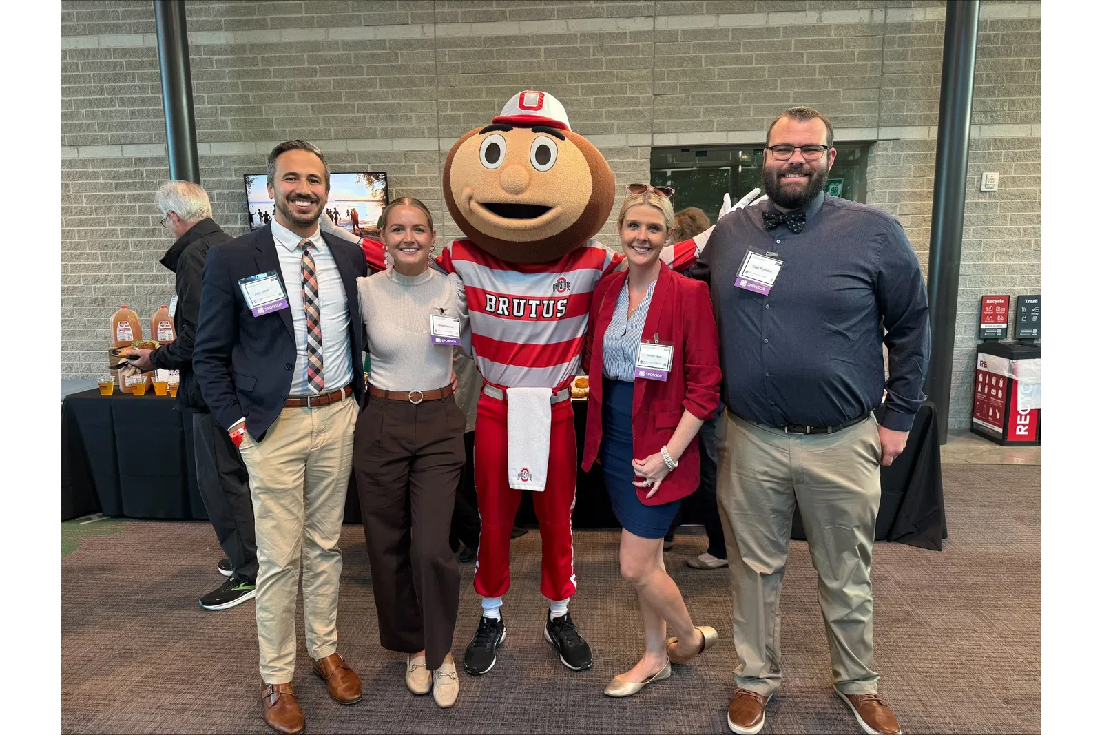 Group of four people posing with Brutus Buckeye mascot at an indoor event with brick walls and tables.