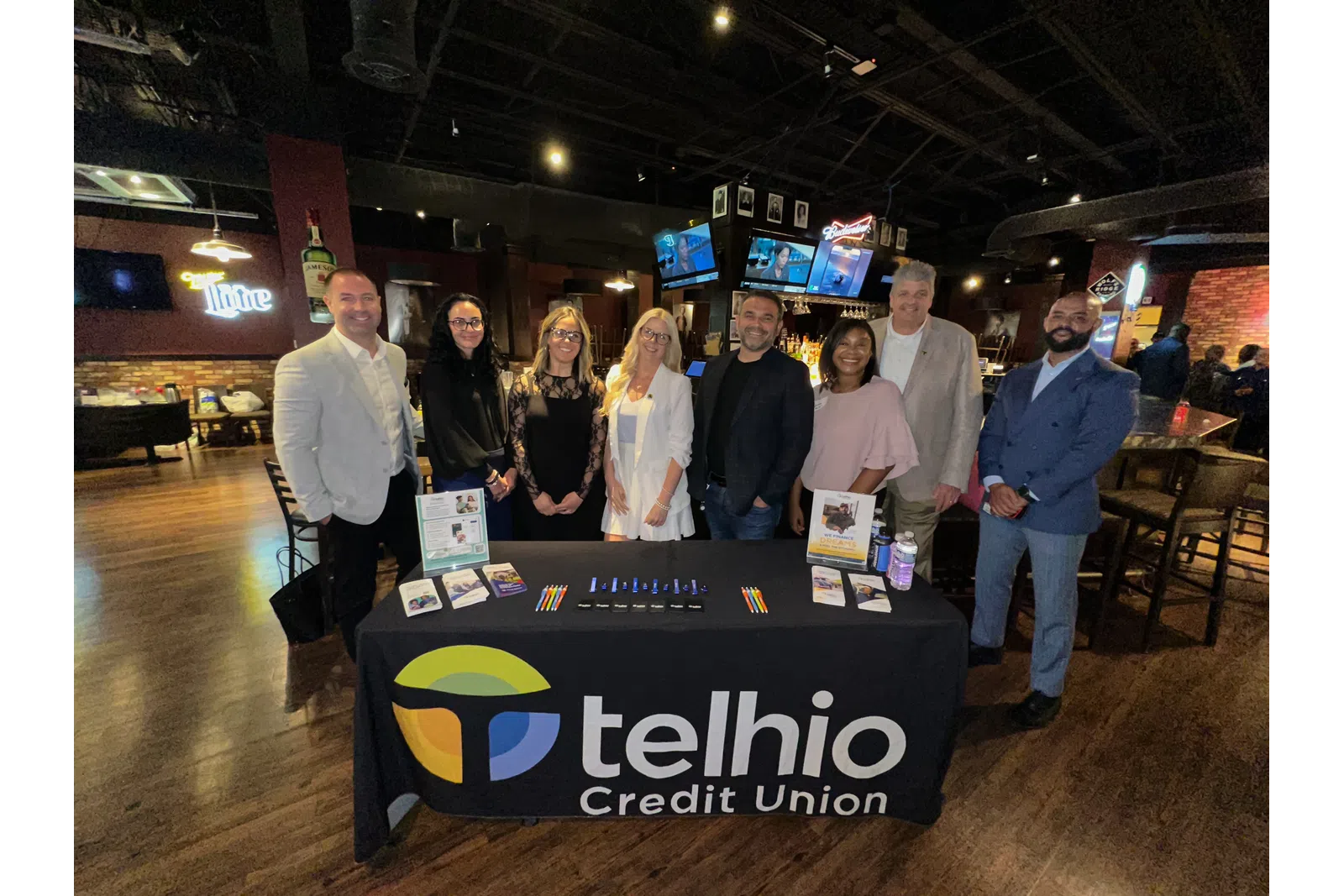Group of professionals standing behind a Telhio Credit Union promotional table in a bar setting.