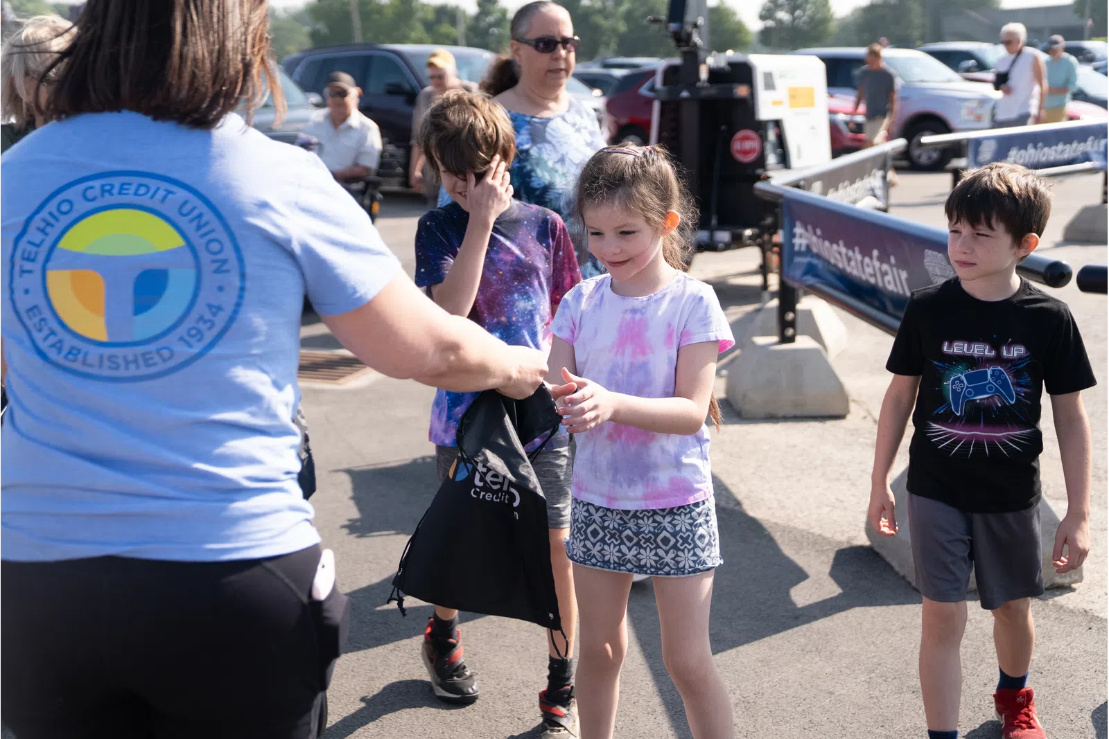 Children receiving giveaway bags from Telhio Credit Union staff during an outdoor event on a sunny day.