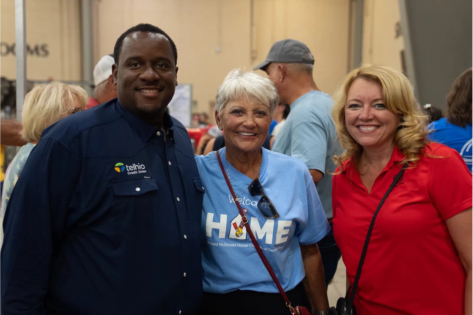 Three smiling adults posing at a community event indoors with casual and branded clothing.