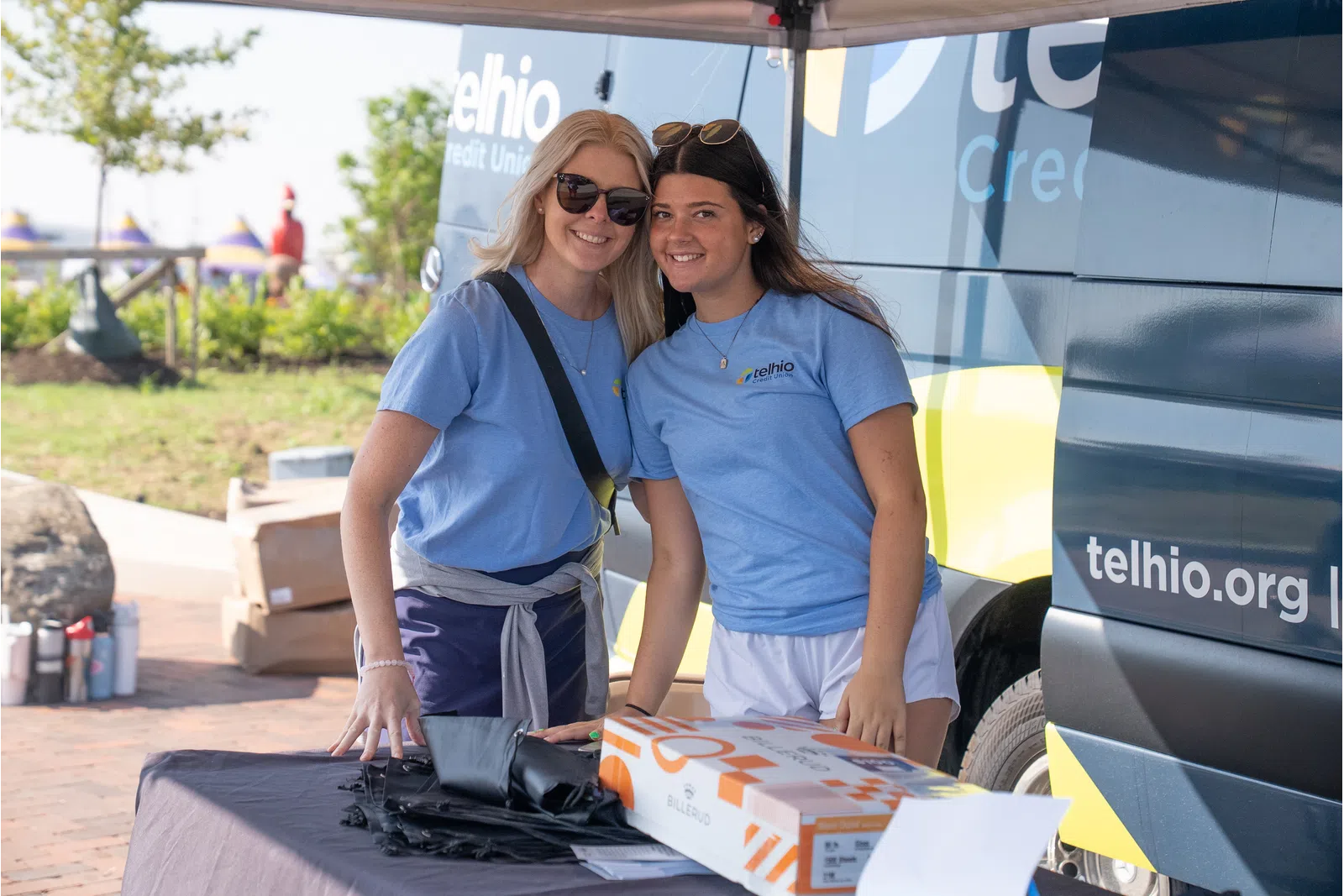 Two women in blue Telhio Credit Union shirts smiling at an outdoor event booth with branded van in background.