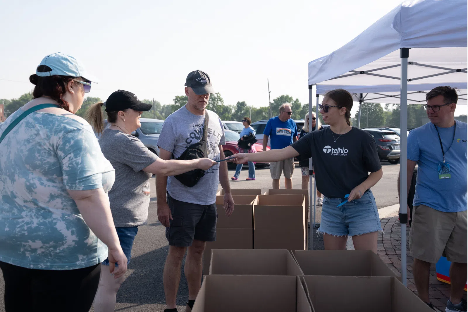 Volunteers distributing items to people at an outdoor event under a white tent with cardboard boxes
