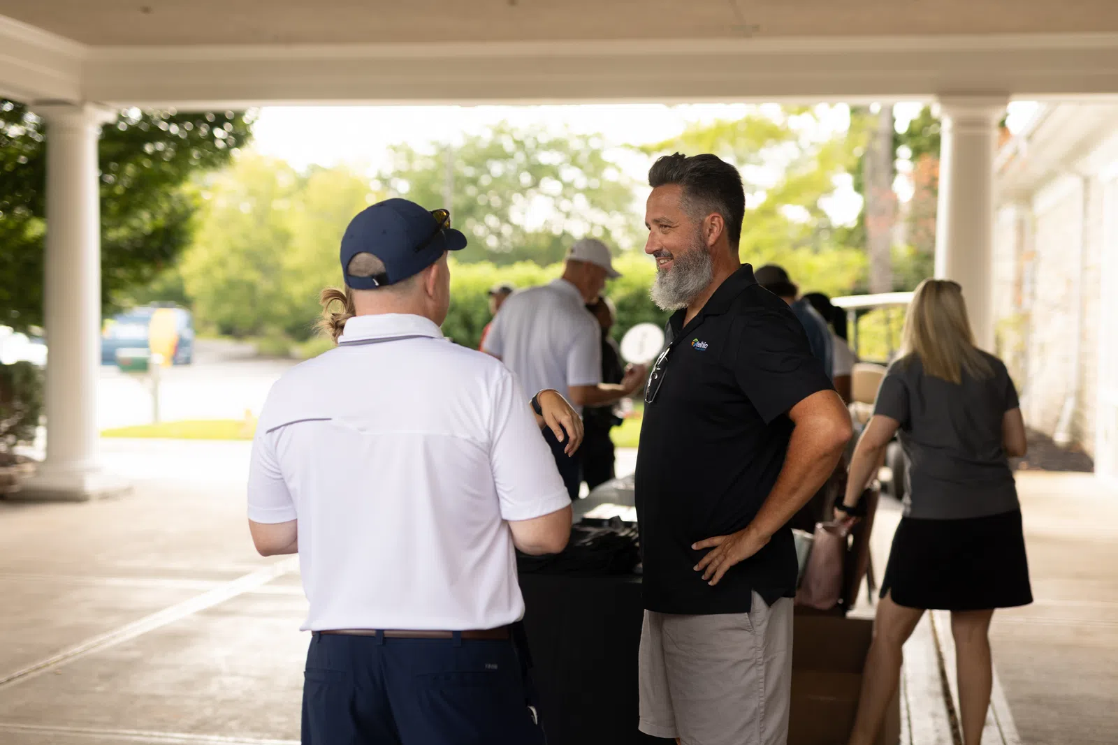 Two men in casual golf attire talking under a covered outdoor area near a table with people and trees in background
