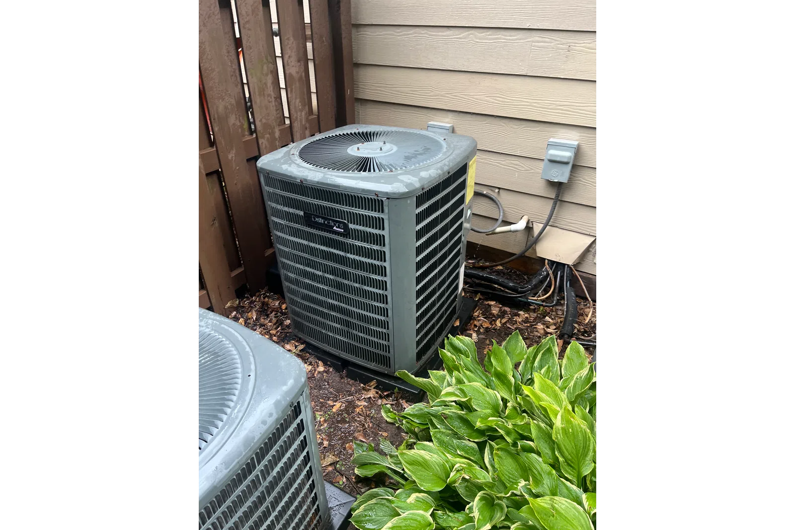 AC Maintenance, Cleaning and Tune-up in Johns Creek by The Air Company of GA.  Two outdoor air conditioning units near a wooden fence and siding with green leafy plants in front.