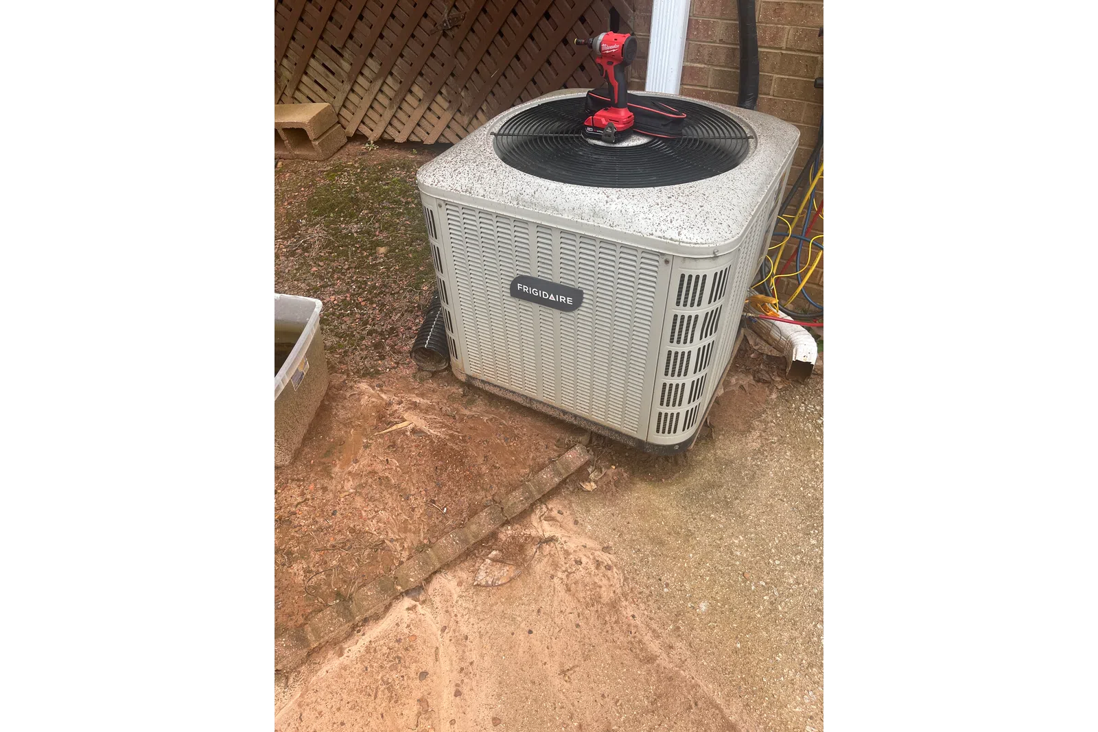 AC Preventative Maintenance in Tucker, Atlanta by The Air Company of GA. Outdoor Frigidaire air conditioner unit with a red Milwaukee drill on top beside a brick wall and garden area.