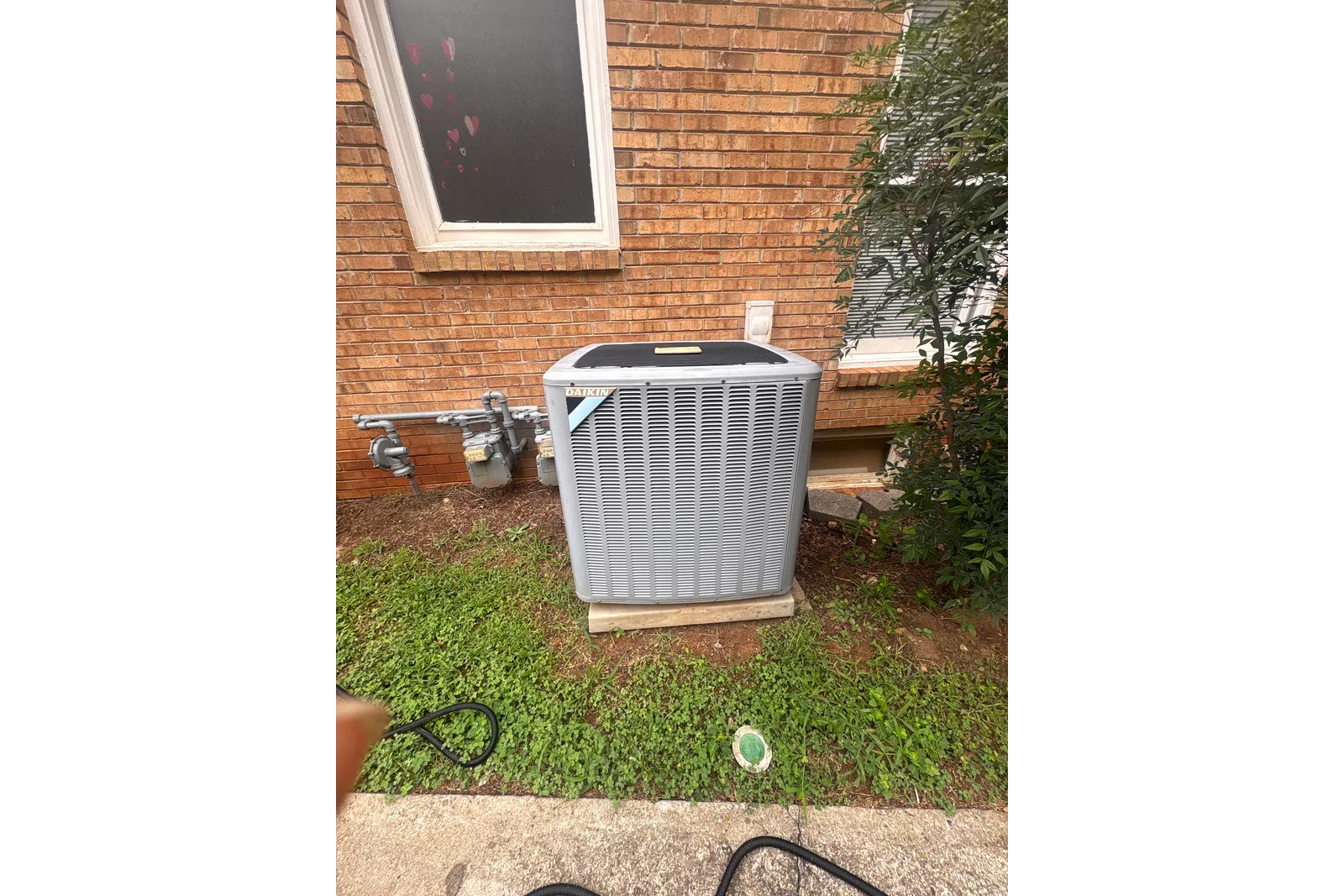 AC Repairs in Tucker Atlanta by The Air Company of GA. Outdoor Daikin air conditioning unit installed beside brick house wall with grass and shrubbery.
