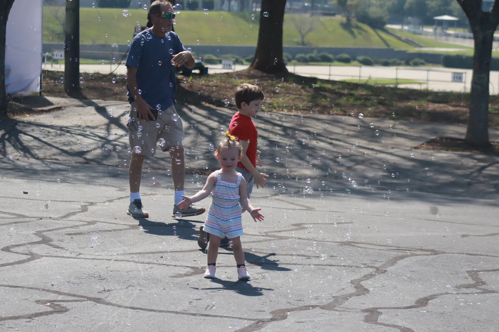 Child in striped romper playing with bubbles outdoors on sunny day, accompanied by boy and adult male.