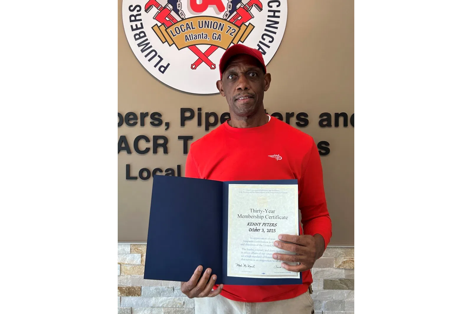 Man in red shirt and cap holding a certification in front of a plumbers and service technicians union sign.