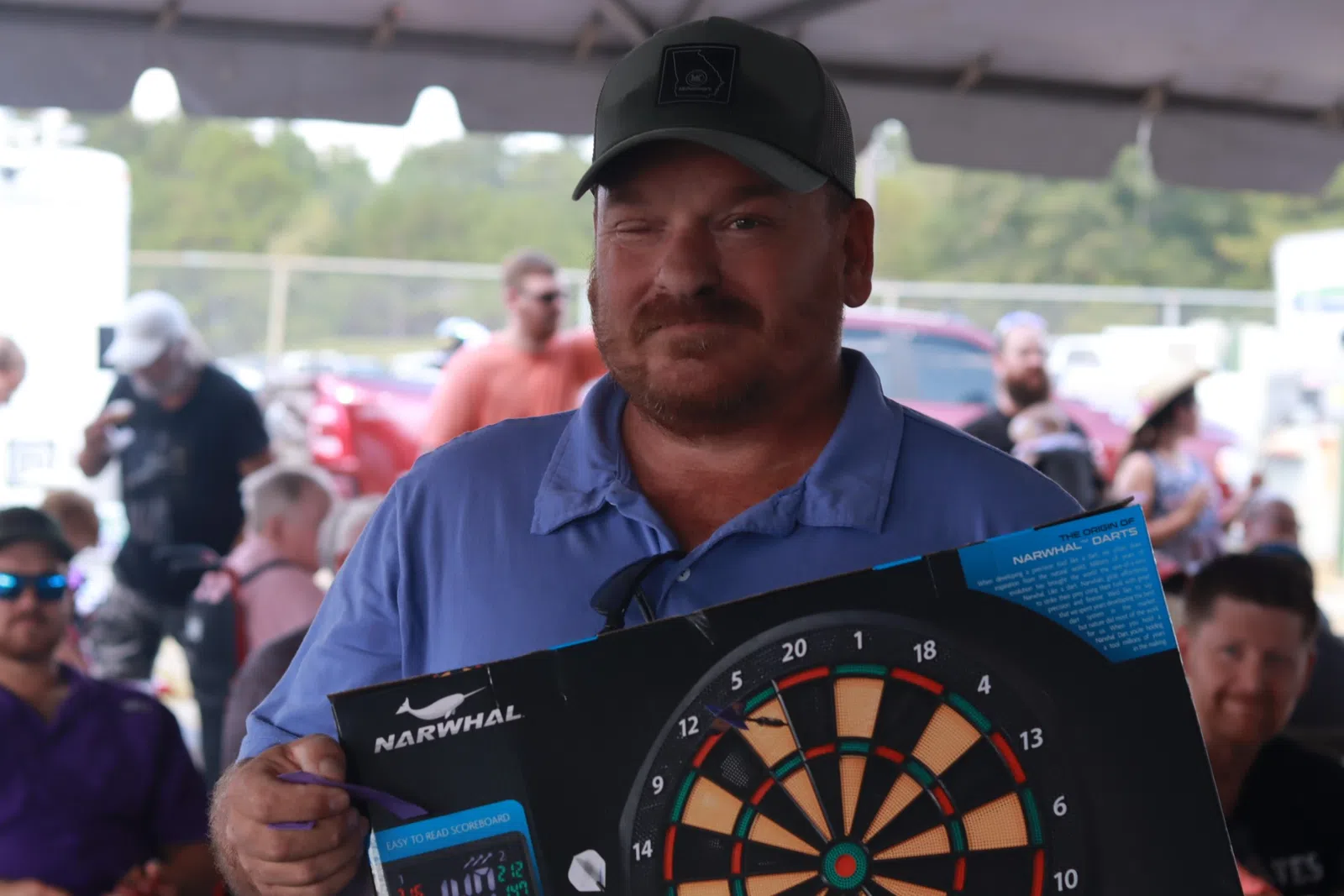 Man in blue shirt and cap holding a Narwhal dartboard with a crowd in background at outdoor event.
