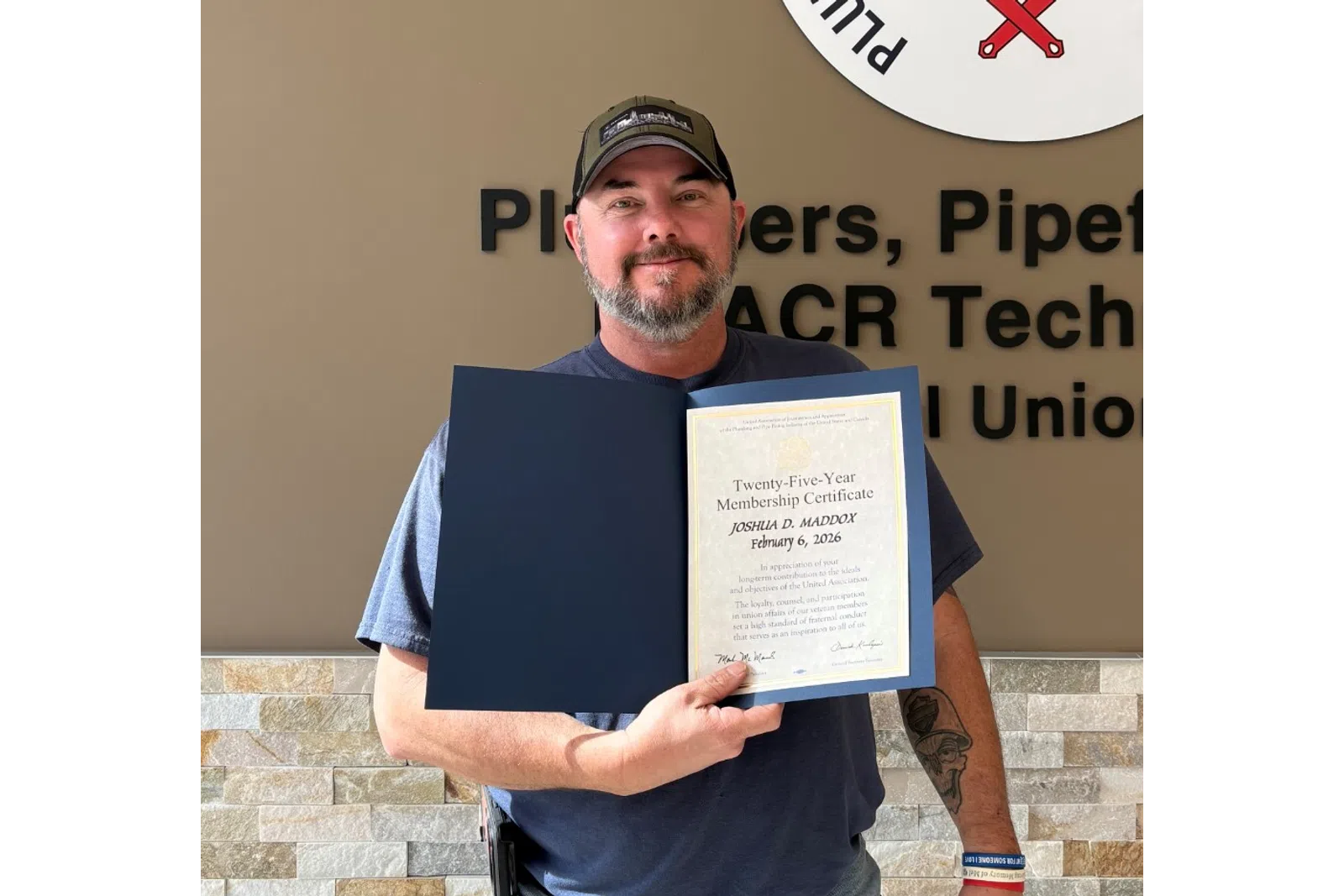 Man in cap holding certificate standing by Plumbers Pipefitters Service Technicians Union logo wall