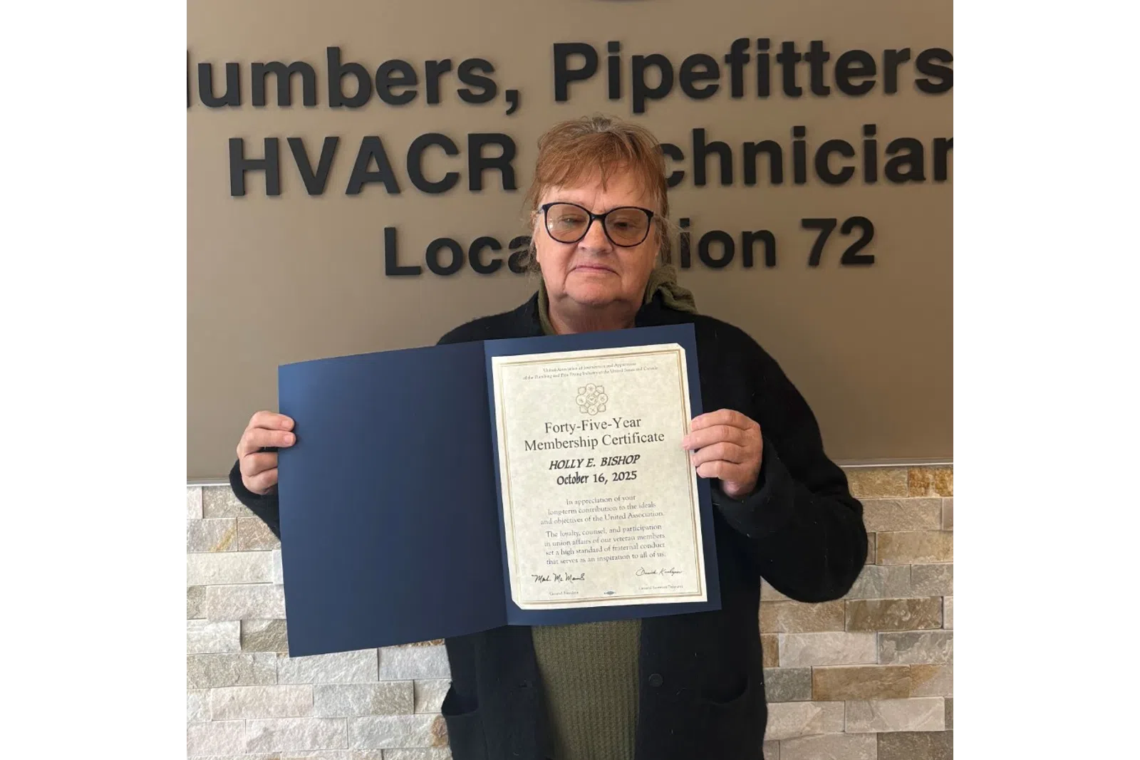 Person holding a certificate standing next to a Plumbers and Pipefitters Union Local 72 sign on a beige wall.