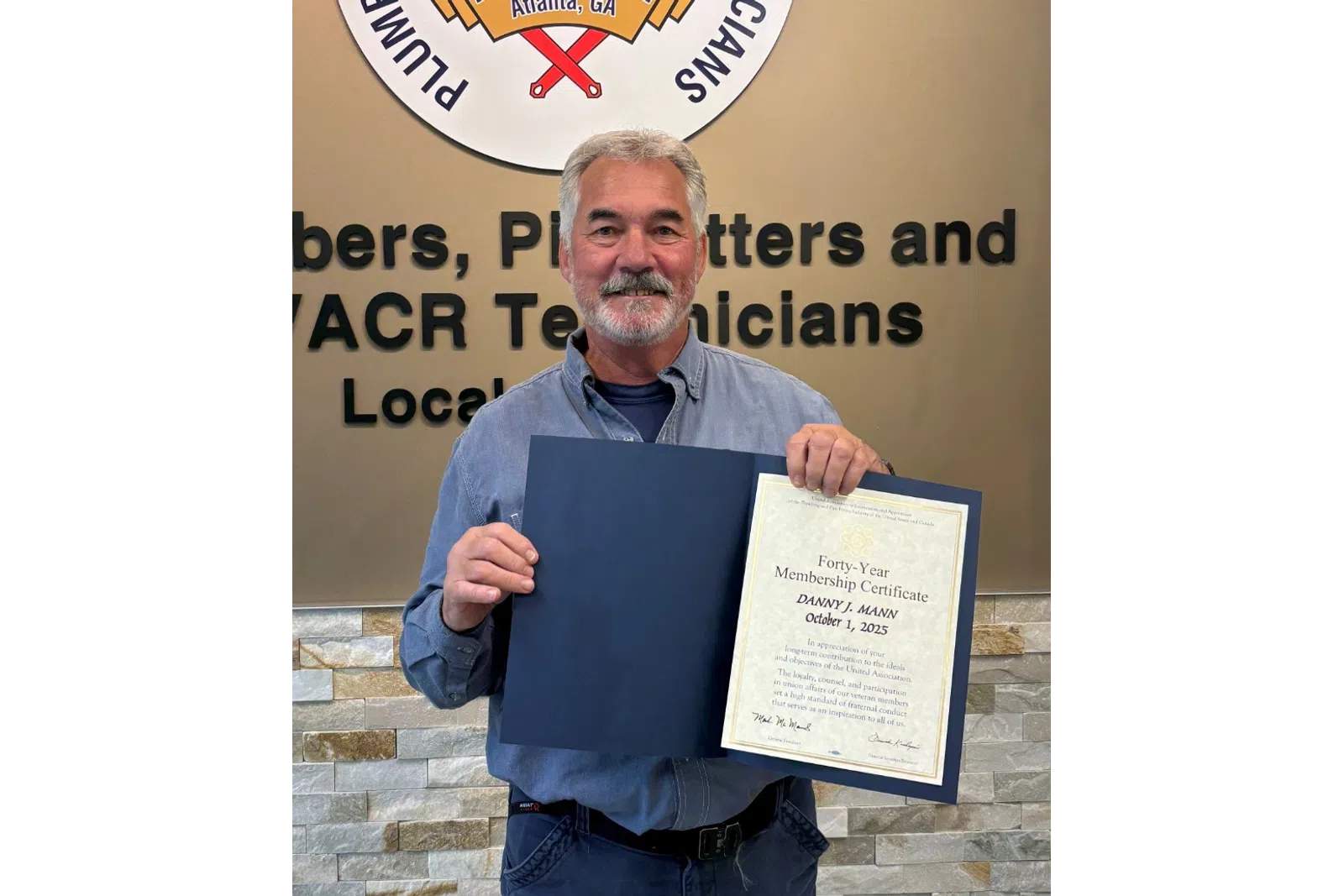 Man holding a certificate standing next to Plumbers and Pipefitters Local Union 72 logo on wall