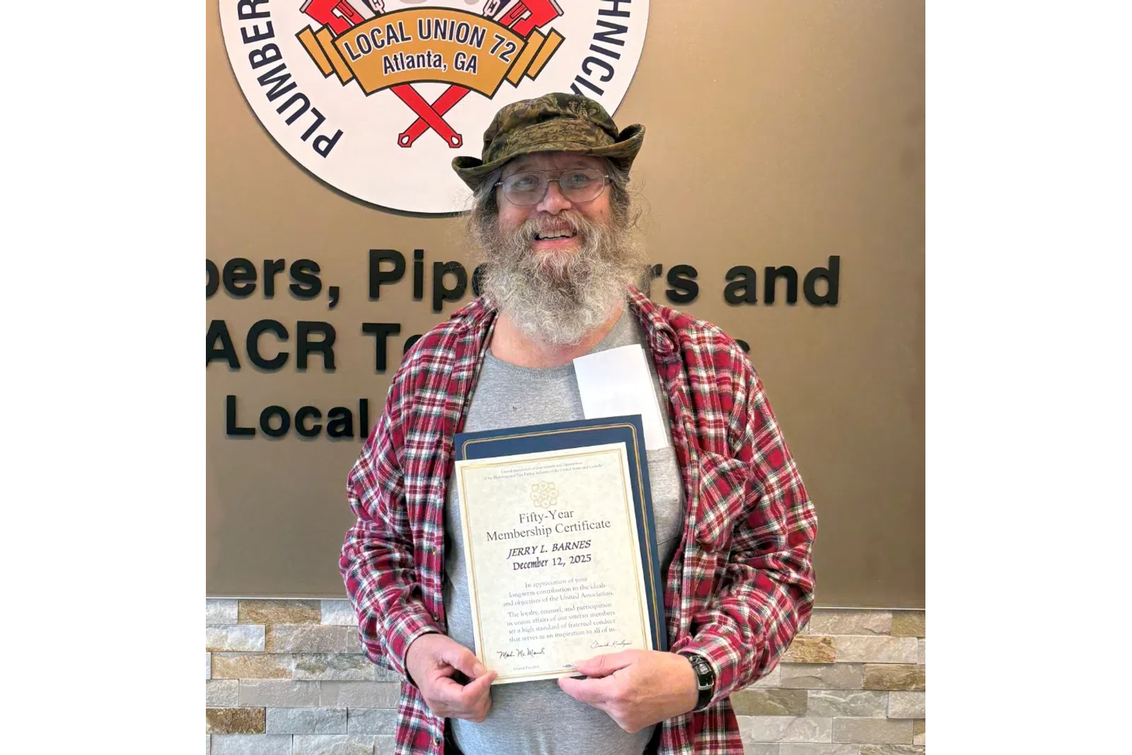 Man in a red plaid shirt holding a certificate standing next to a Plumbers service union sign on a wall.