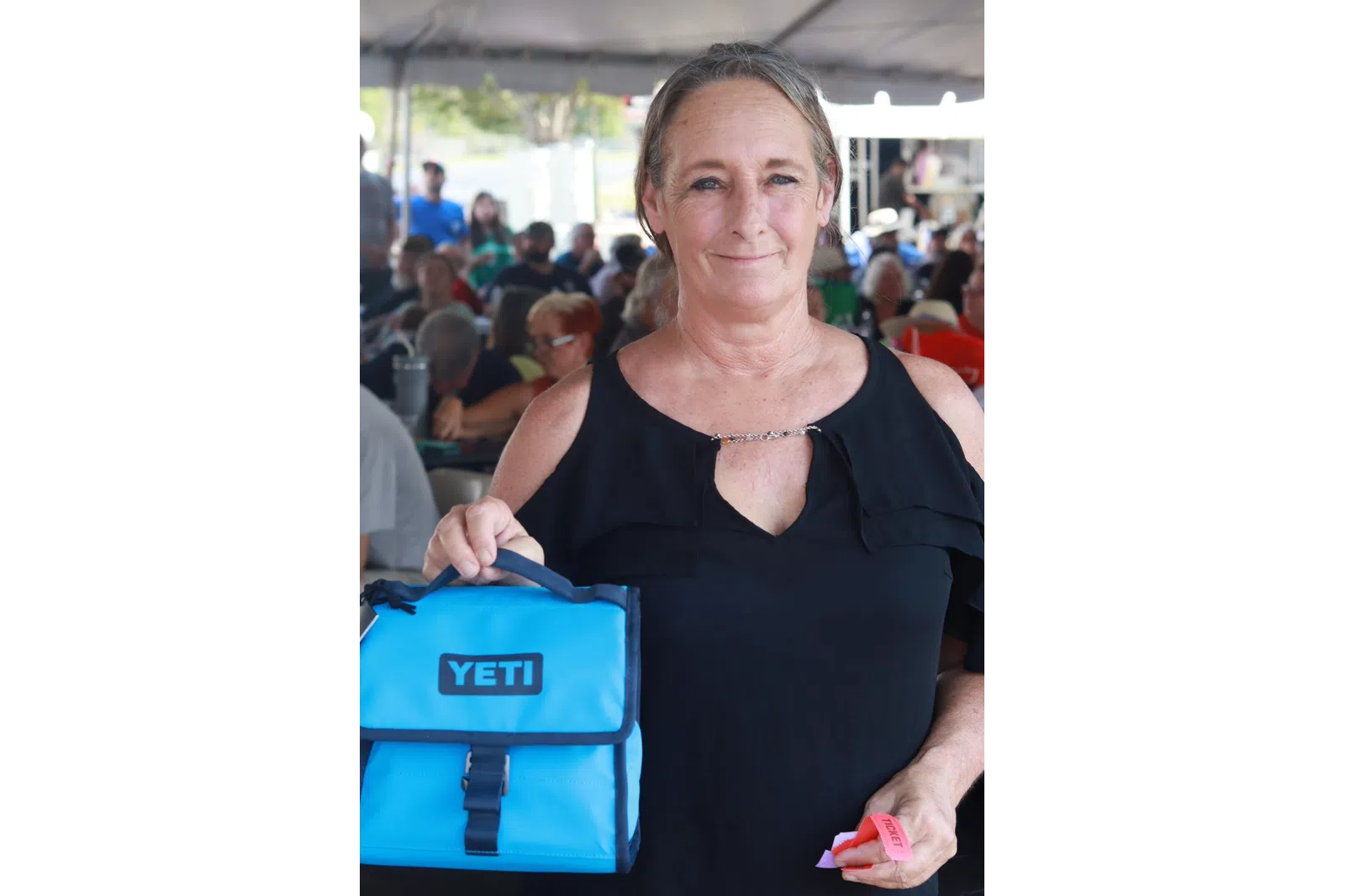 Woman holding a blue YETI cooler bag and event tickets at an outdoor gathering under a tent.
