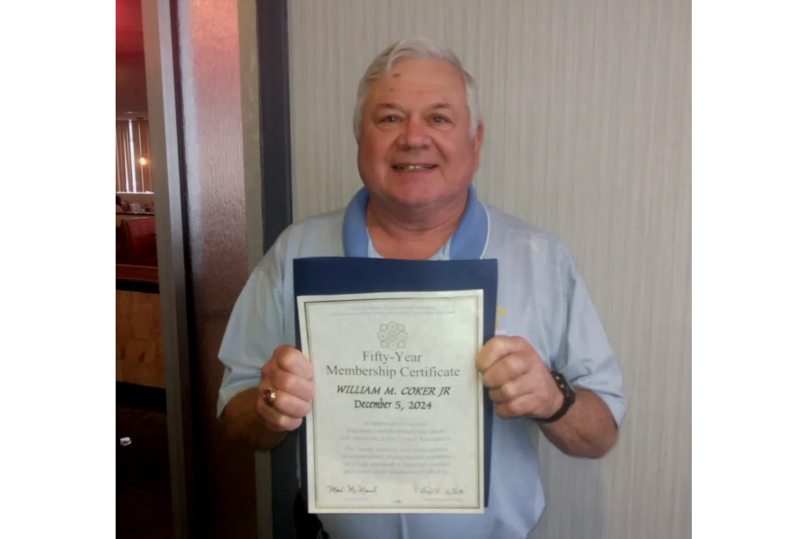 Man holding a fifty-year membership certificate with a smile, standing indoors against a neutral wall.