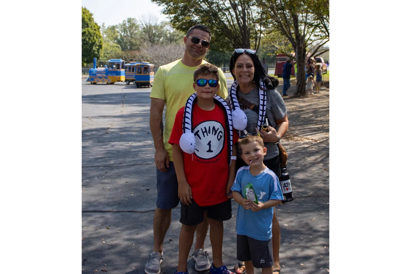 Smiling family of four posing outdoors on a sunny day with trees and a small train in the background.