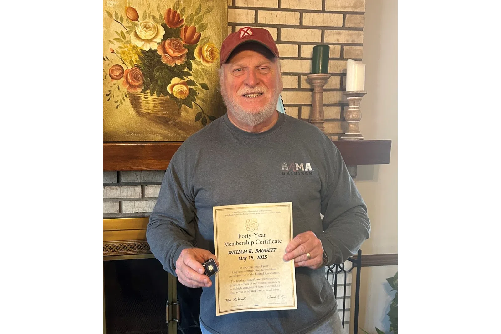 Smiling man holding a certificate standing by a brick fireplace with floral painting and plants.
