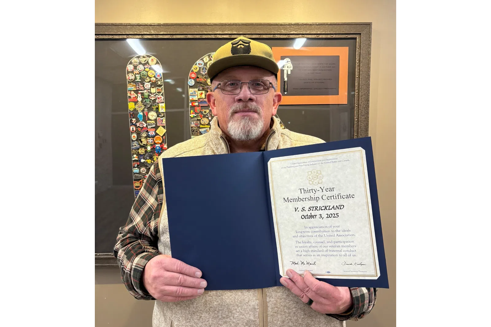 Man with beard and glasses holding a thirty-year membership certificate in front of a framed pin collection.
