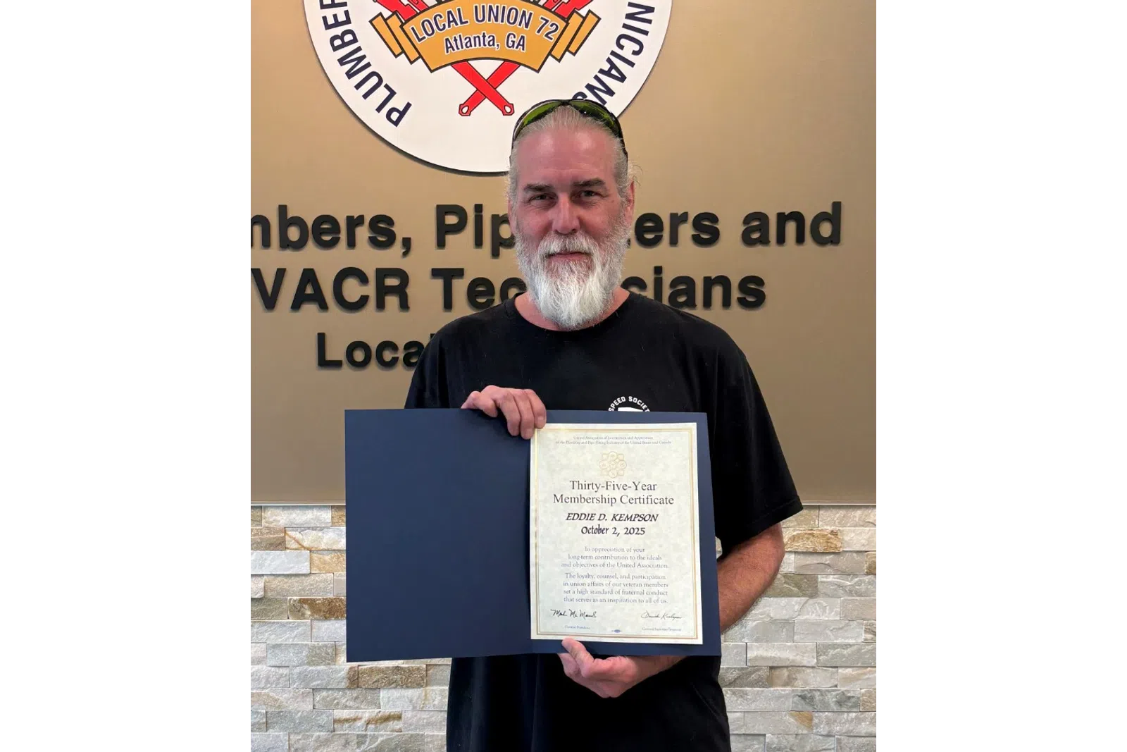 Man with gray beard holding a certificate standing in front of a Plumbers and Pipefitters union sign and wall