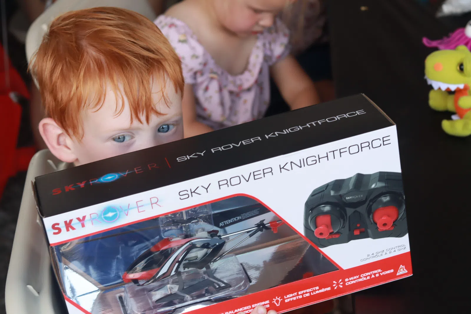 Young red-haired child holding a Sky Rover Knightforce remote control helicopter box with another child in background.